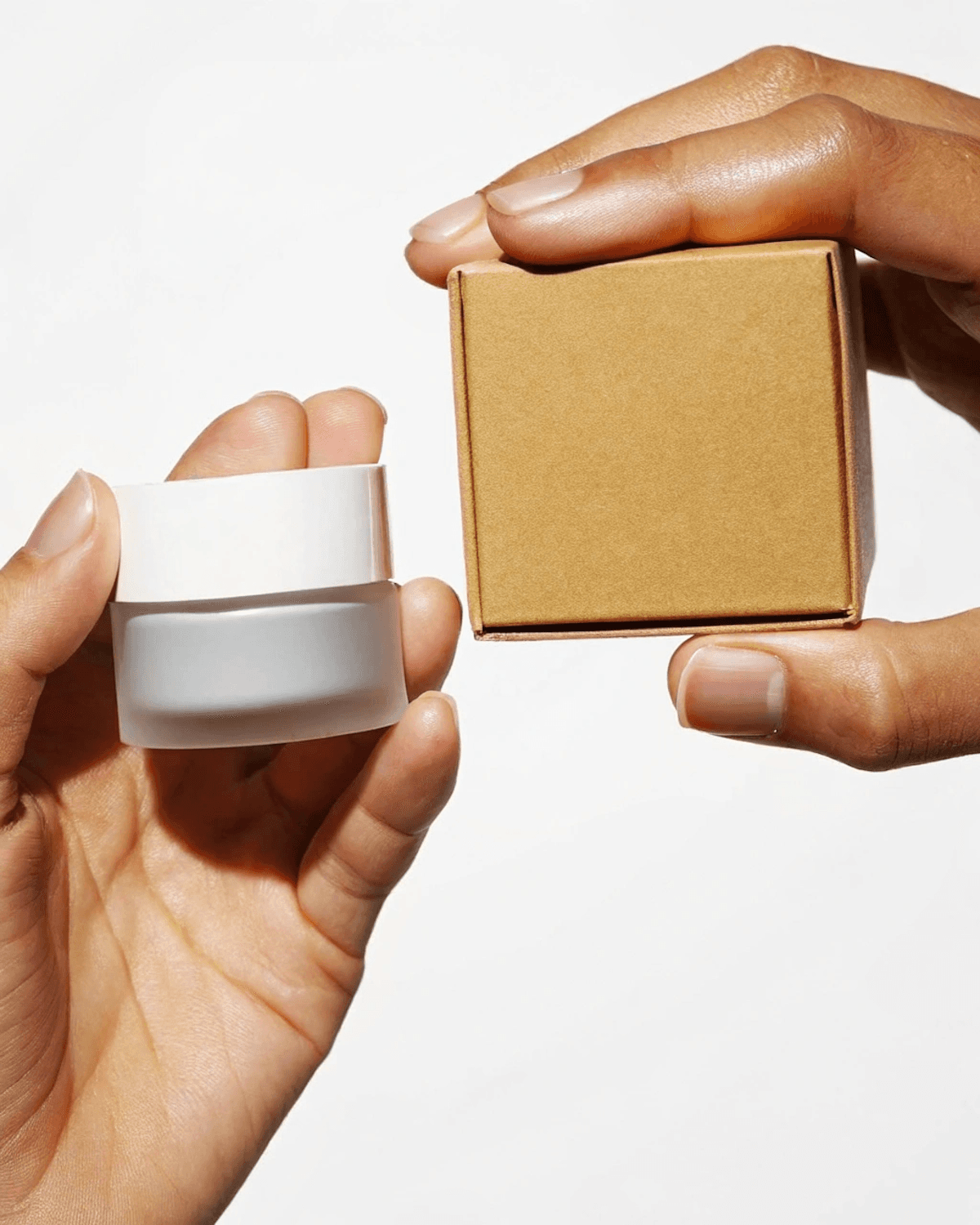 Frosted glass jar of moisturizer held next to its kraft paper gift box