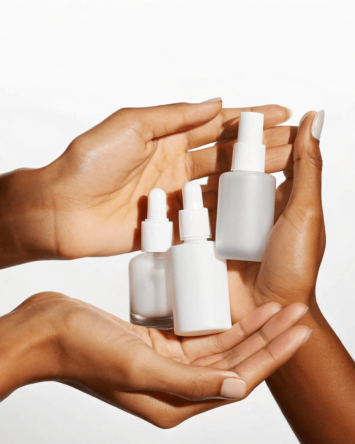 Hands holding three white skincare bottles: two dropper serums and a spray mist